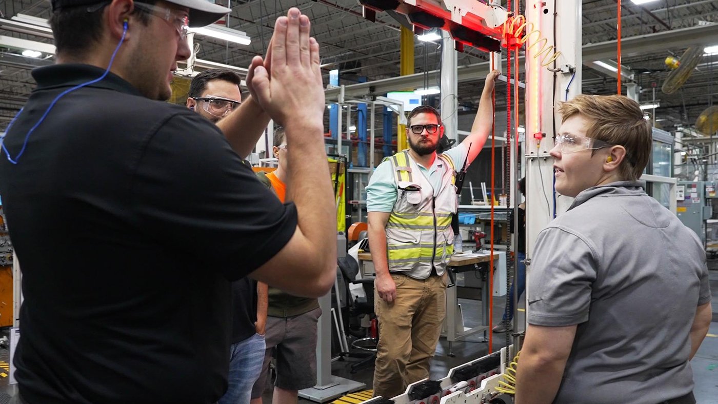 Workers gather on a factory floor while one person explains equipment at a workstation.