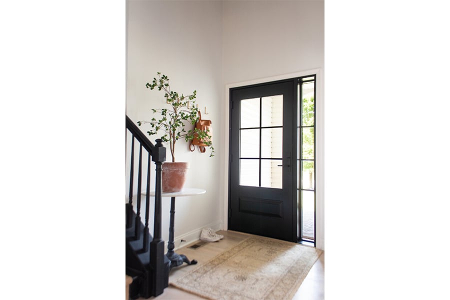 An interior shot of an entryway with a rug, table with a potted plant, and black front door whose window has colonial grilles and a sidelight to match.