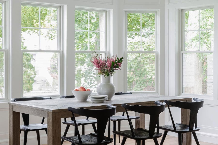 A breakfast room with a dining table and chairs is flooded with light from the three walls of windows with modified colonial grilles.