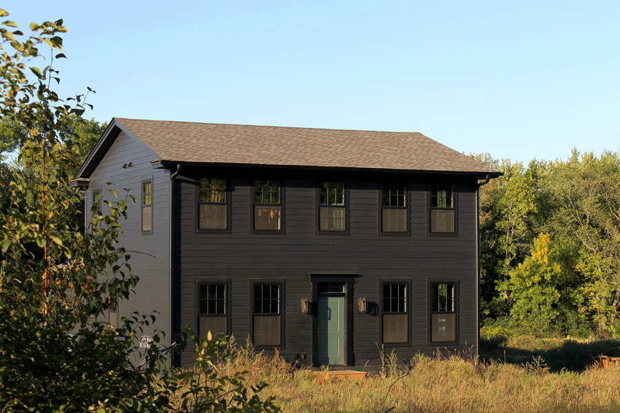 A black home with black windows and a green front door stands out against a landscape of prairie and woods.