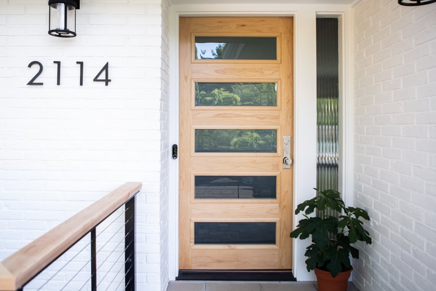 A modern front door with a light-colored wood exterior and horizontal glass panels popping against the white brick walls of a Mid-Century Modern home.